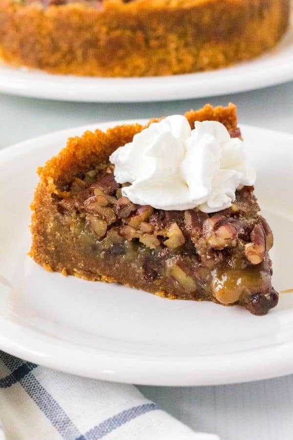 A slice of Instant Pot pecan pie with chocolate chips topped with whipped cream on a white plate, with the rest of the pie in the background.