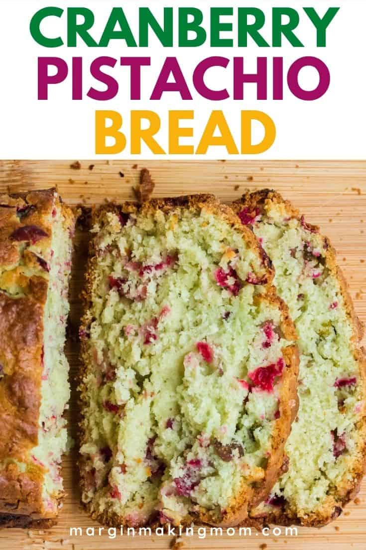 a loaf of pistachio cranberry bread sliced on a cutting board, with two slices laying open.