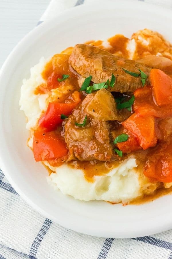 Pressure cooker Swiss Steak served over a bed of mashed potatoes on a white plate, set on a blue and white checked napkin.