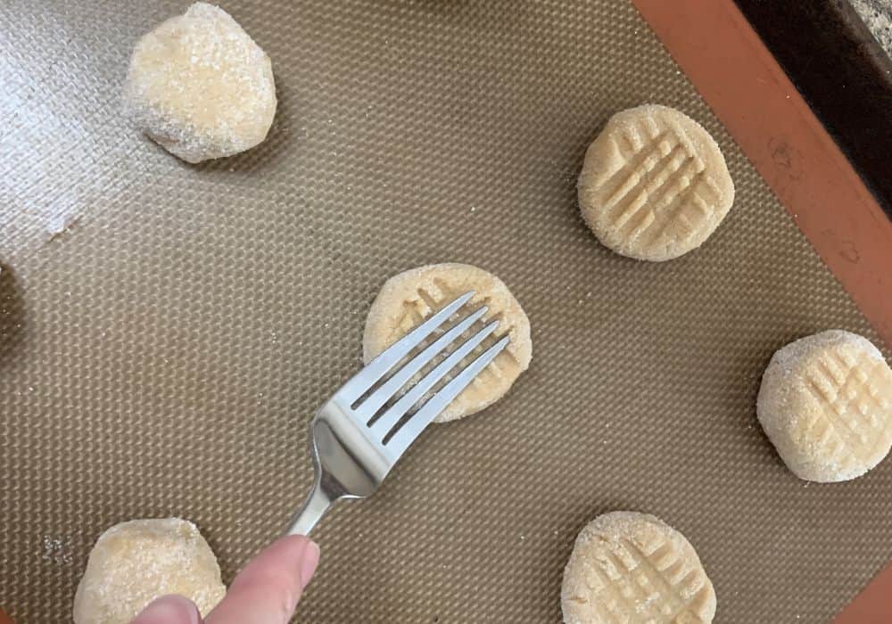 tines of a fork pressing a criss-cross pattern into the pancake mix peanut butter cookies on a baking sheet