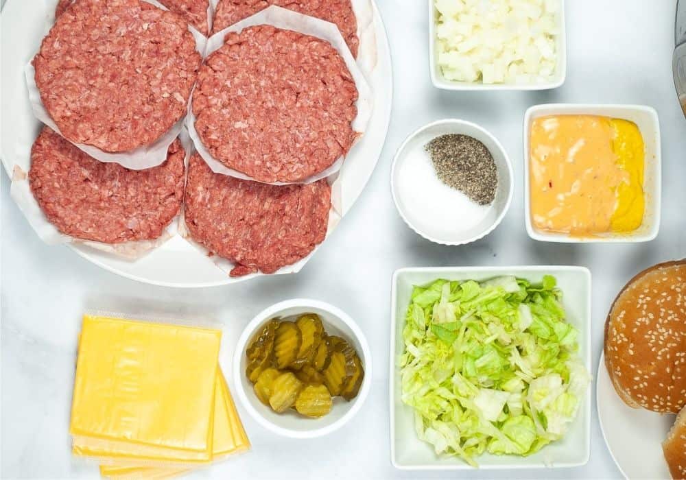 a plate of raw hamburger patties for making burgers in the Instant Pot, plus various toppings including cheese, pickles, lettuce, salt, pepper, dressing, onions, and buns.