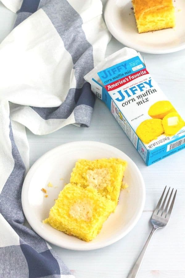 Jiffy cornbread box next to two white plates with servings of cornbread on them, alongside a blue and white cloth napkin and a fork.