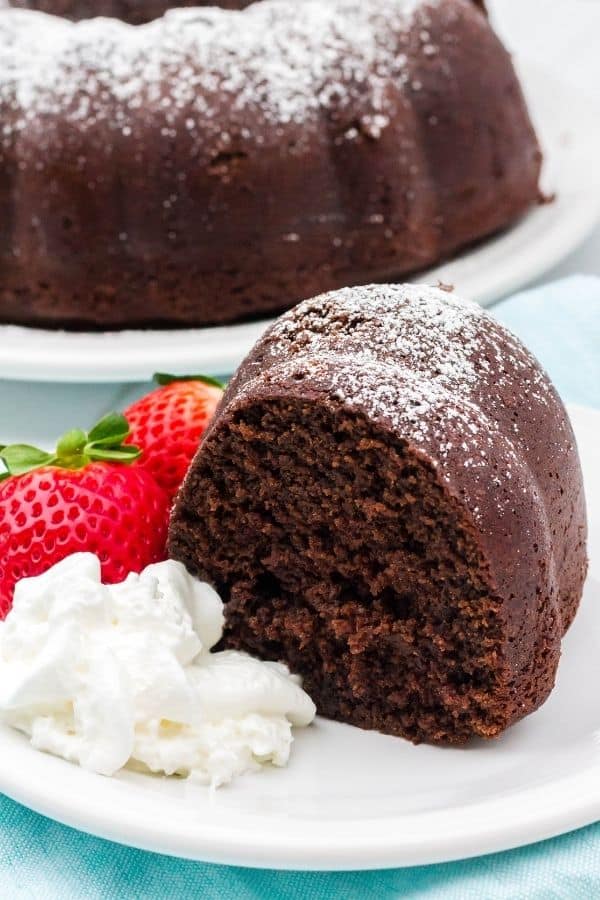 Strawberries, whipped cream, and a piece of chocolate bundt cake on a white plate, set over a light blue napkin.