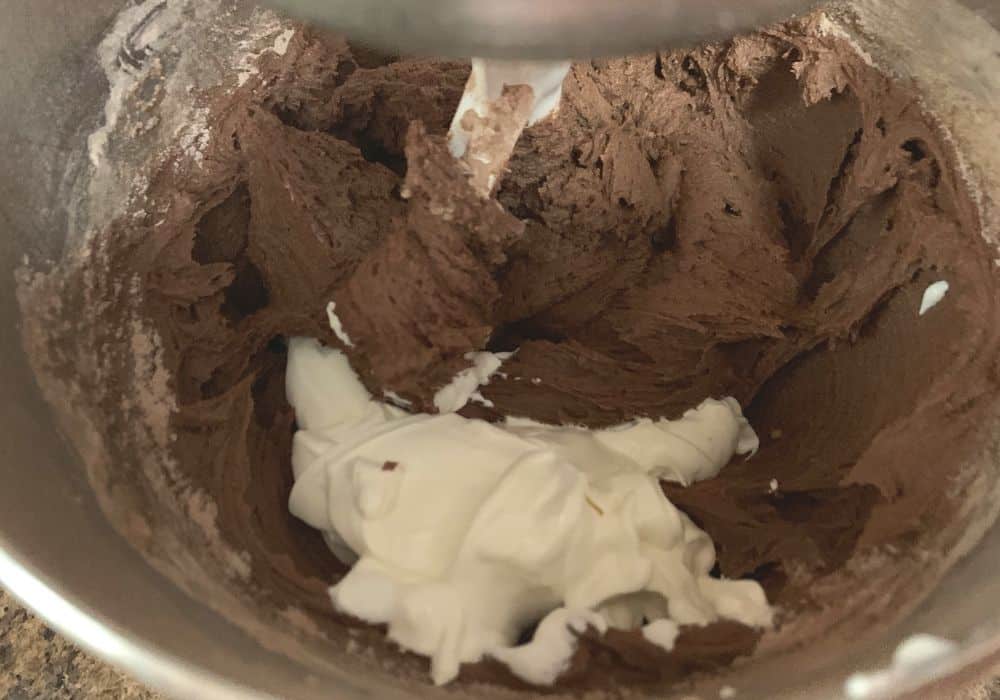 Sour cream added to thick batter in a mixing bowl for making chocolate bundt cake.