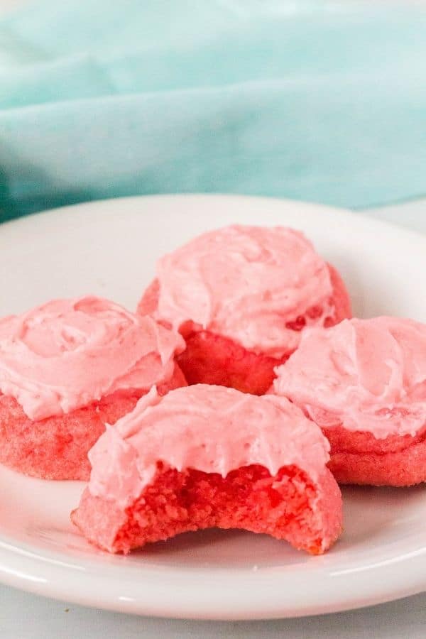 soft strawberry sugar cookies made with Jello and frosted with pink icing are on a white plate. One cookie has a bite taken out of it.