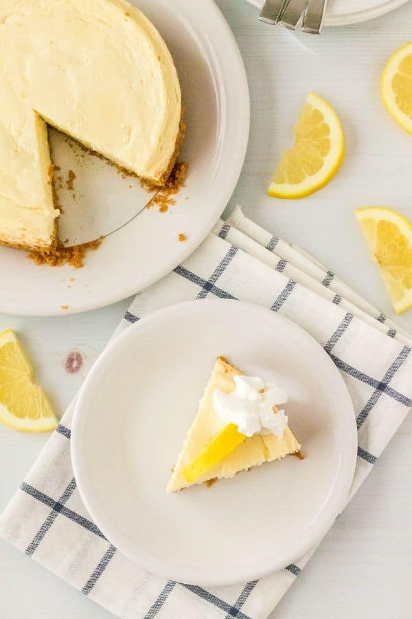 slice of Instant Pot lemon cheesecake on a white plate, atop a blue and white napkin, with the remaining cheesecake behind it and lemon garnishes surrounding the plate.