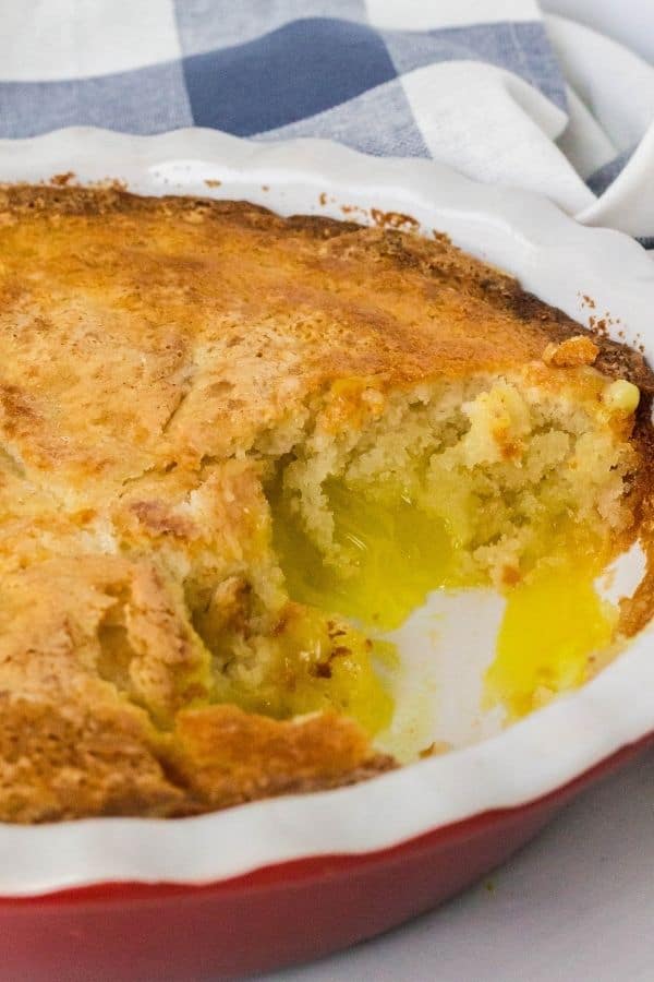 red deep dish pie plate filled with lemon cobbler, with a scoop removed showing the custard filling under the cake-like topping