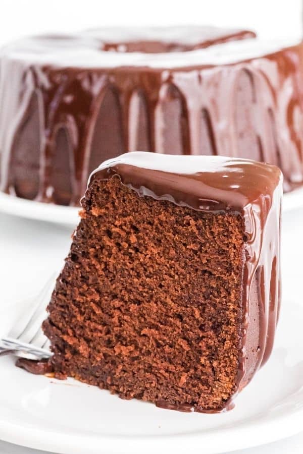 A slice of chocolate sour cream cake on a white plate, with the remaining cake in the background