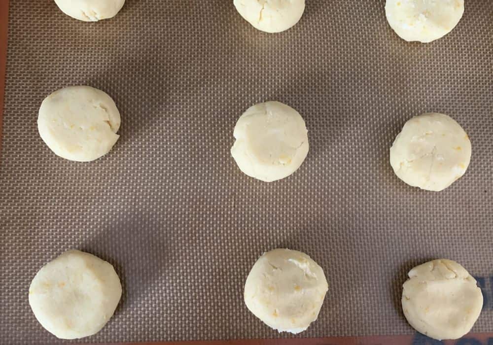 several rounds of slightly flattened lemon cookie dough on a lined baking sheet, ready to be baked