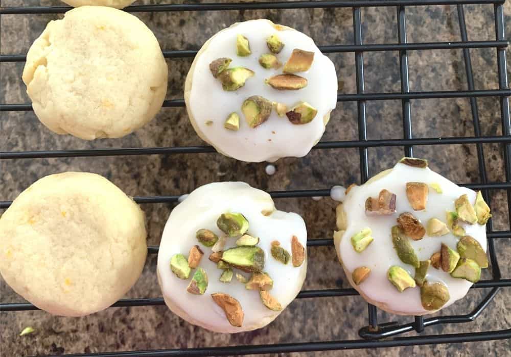 lemon cookies on a cooling rack. 3 of the cookies have been dipped in the lemon icing and topped with chopped pistachios.