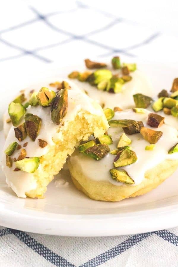 a few frosted lemon cookies topped with pistachios on a white plate, with a bite taken out of one cookie showing its tender interior