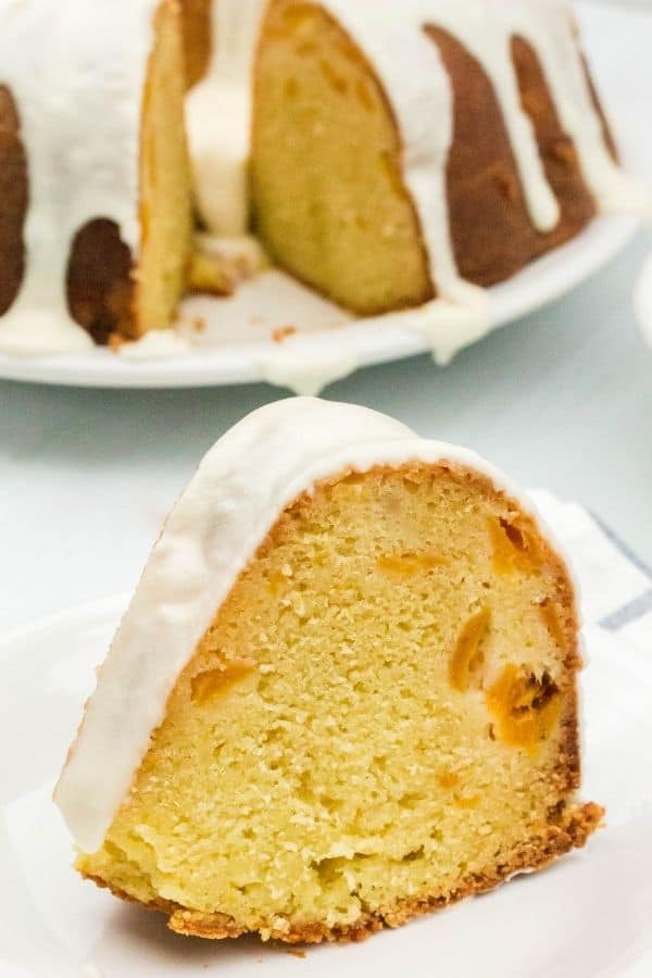 slice of sour cream pound cake, dotted with peaches, topped with glaze, served on a white dessert plate. Remaining bundt cake is in the background on a serving platter.