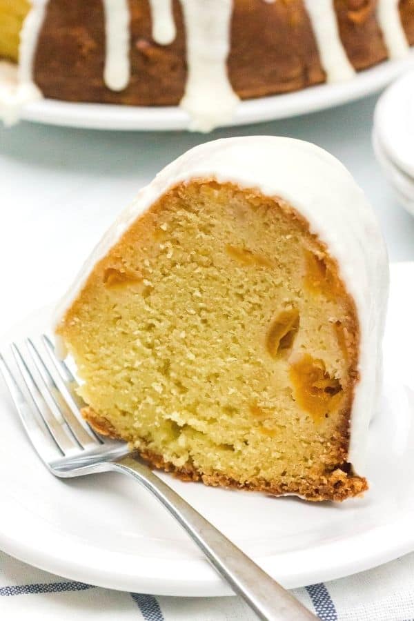 moist and tender sour cream pound cake with peaches on a white plate, with a fork on the plate and the remainder of the bundt cake in the background.