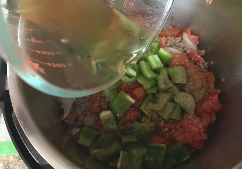 green peppers, tomatoes, ground beef, and onions in the Instant Pot, with beef broth being poured into the pot.