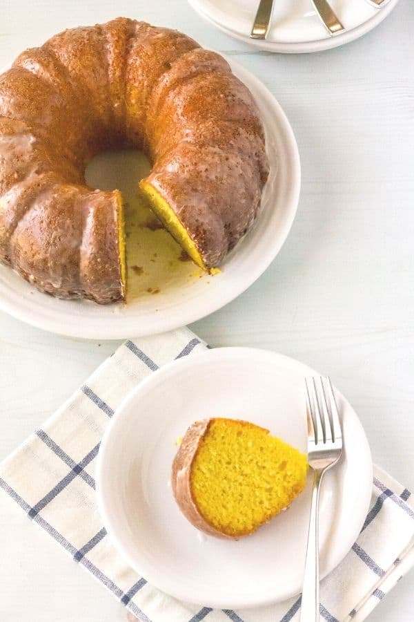 A slice of apricot nectar cake on a white plate with a fork next to it, and the serving plate with the remainder of the whole cake behind it.