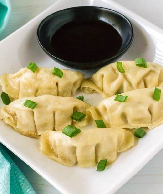 square-shaped white plate serving five Instant Pot frozen dumplings, topped with green onions and served with a side of soy sauce for dipping.
