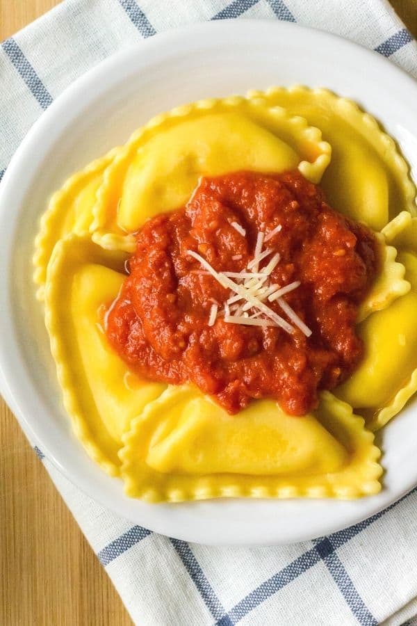overhead view of a white plate atop a blue and white napkin. On the plate is frozen cheese ravioli that was cooked in the Instant Pot, then topped with a tomato sauce and shredded cheese.