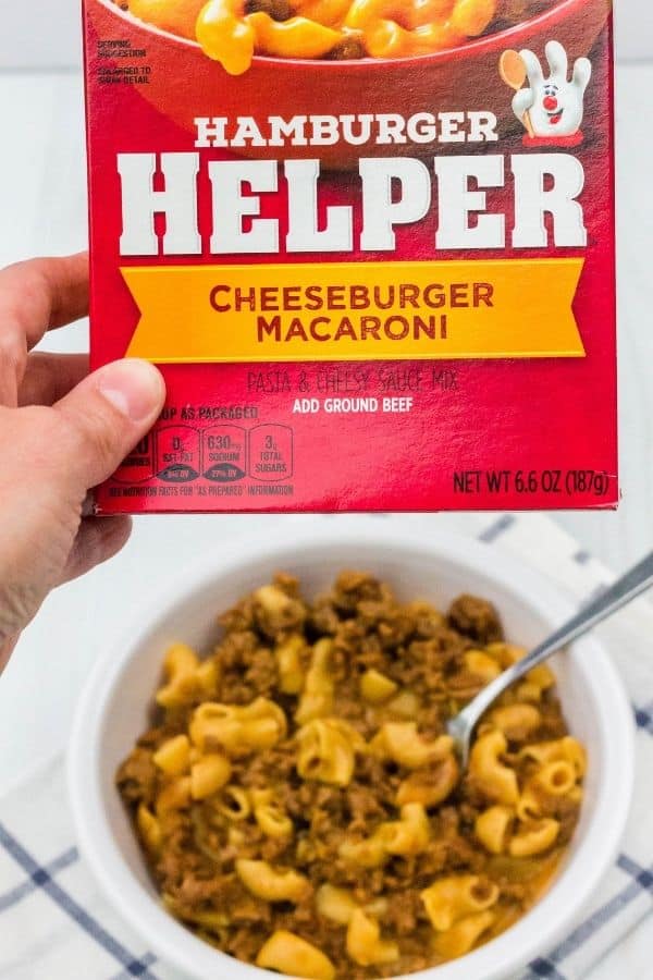 a woman's hand holding a box of Hamburger Helper cheeseburger macaroni in the foreground, with a white bowl of cooked Instant Pot Hamburger Helper below in the background.