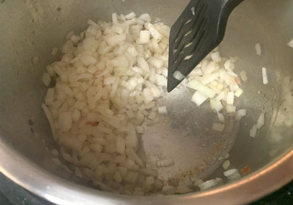 onions being sauteed in bacon grease in the Instant Pot, with a black spatula deglazing the pot