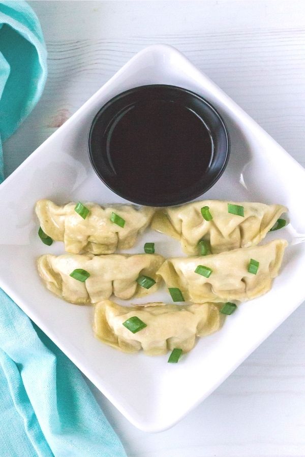 Square white plate with five Asian dumplings on it, topped with green onions, served with a small bowl of soy sauce for dipping.