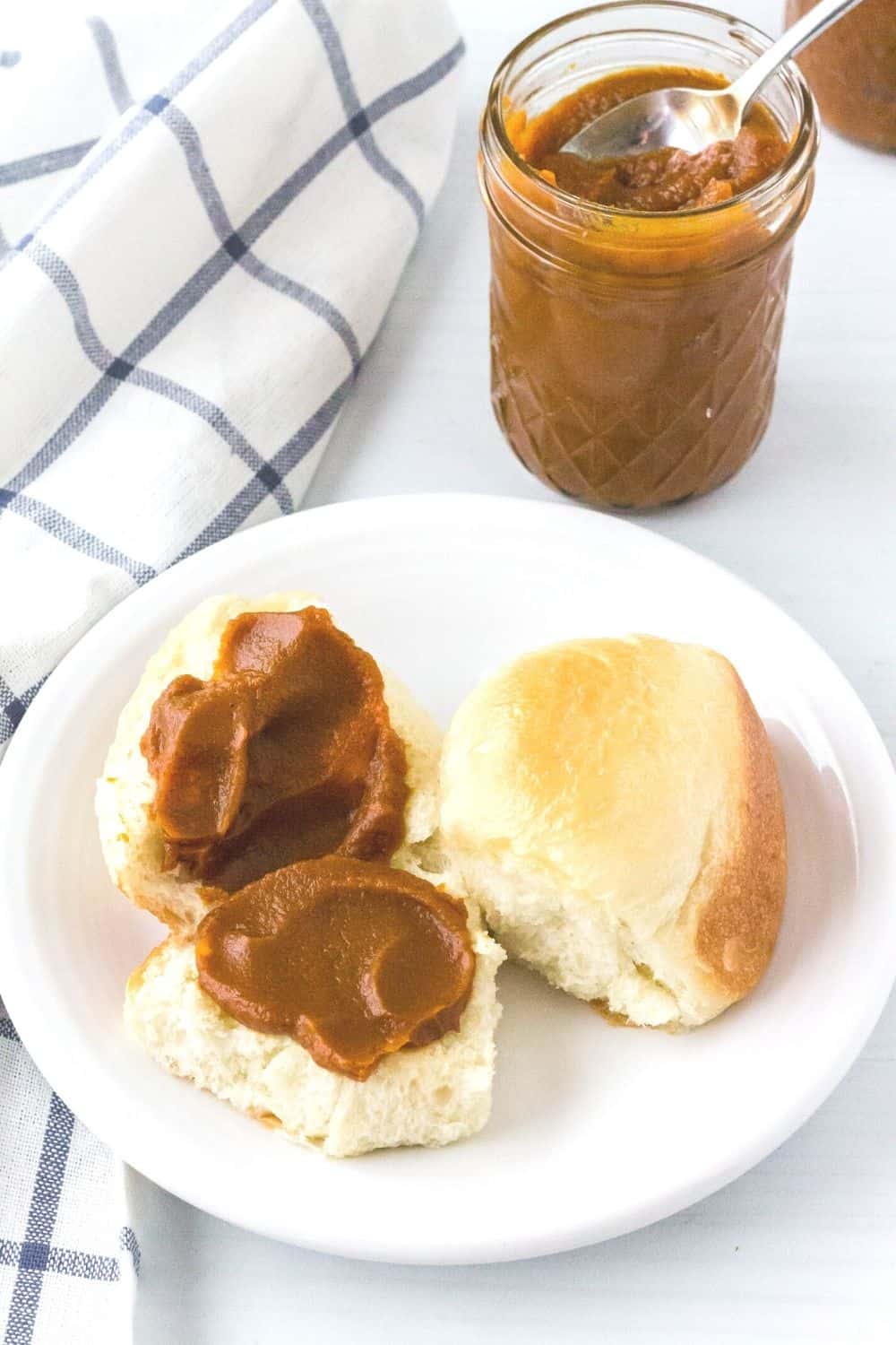 pressure cooker pumpkin butter spread on a halved dinner roll and served on a white plate, with the jar of pumpkin butter in the background.