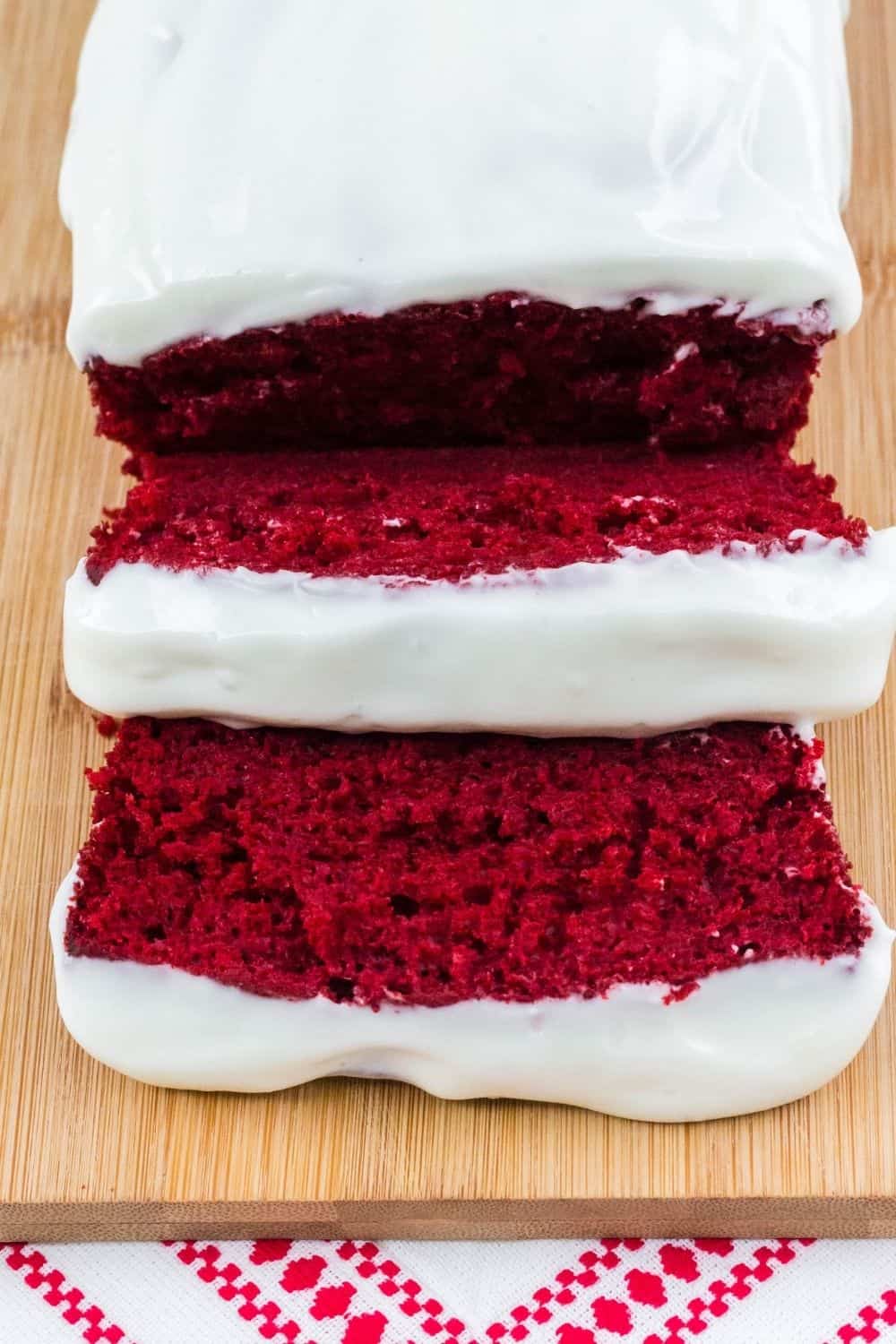 overhead view of a loaf of red velvet cake topped with cream cheese, on a cutting board, sliced.