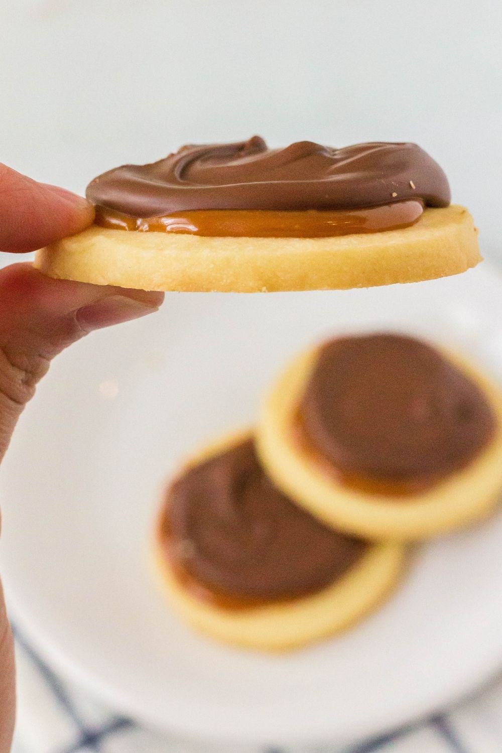 A woman's hand holds a Twix cookie, showing the side view of the cookie, while a plate of cookies rests below.