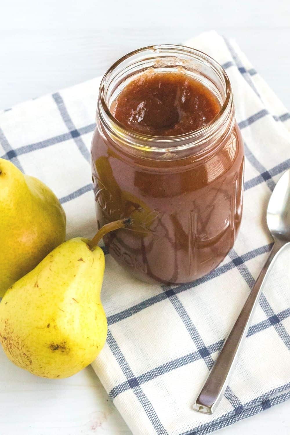 a jar of pear butter on a blue and white checked napkin, with a spoon and two fresh pears next to it.