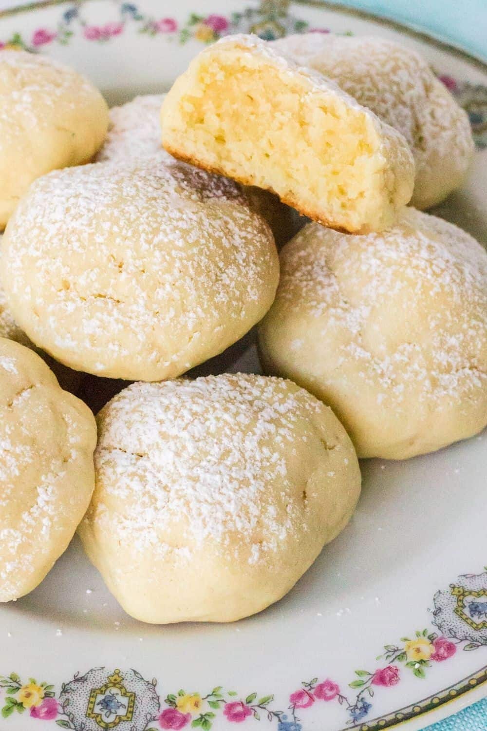 philadelphia cream cheese cookies on a china plate, each dusted with powdered sugar. One cookie has had a bite taken out of it.