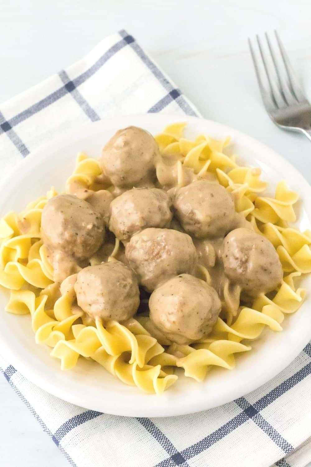 pressure cooker swedish meatballs served over egg noodles on a white plate, resting atop a blue and white napkin with a fork in the background