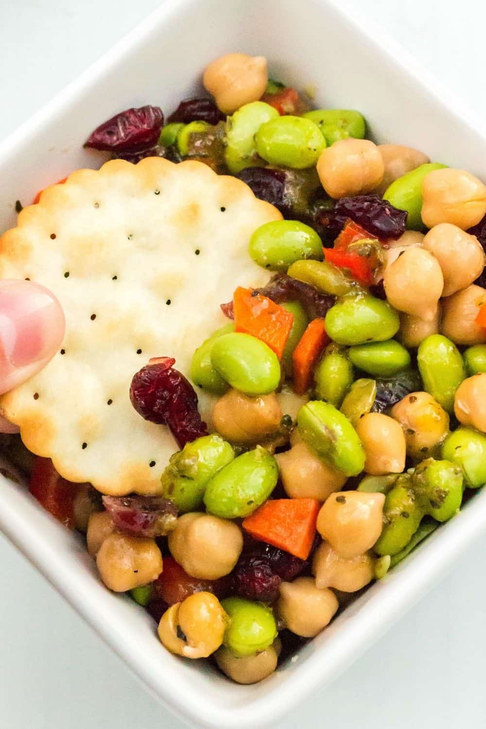 a woman's fingers dip a cracker into a bowl of chickpea and edamame salad