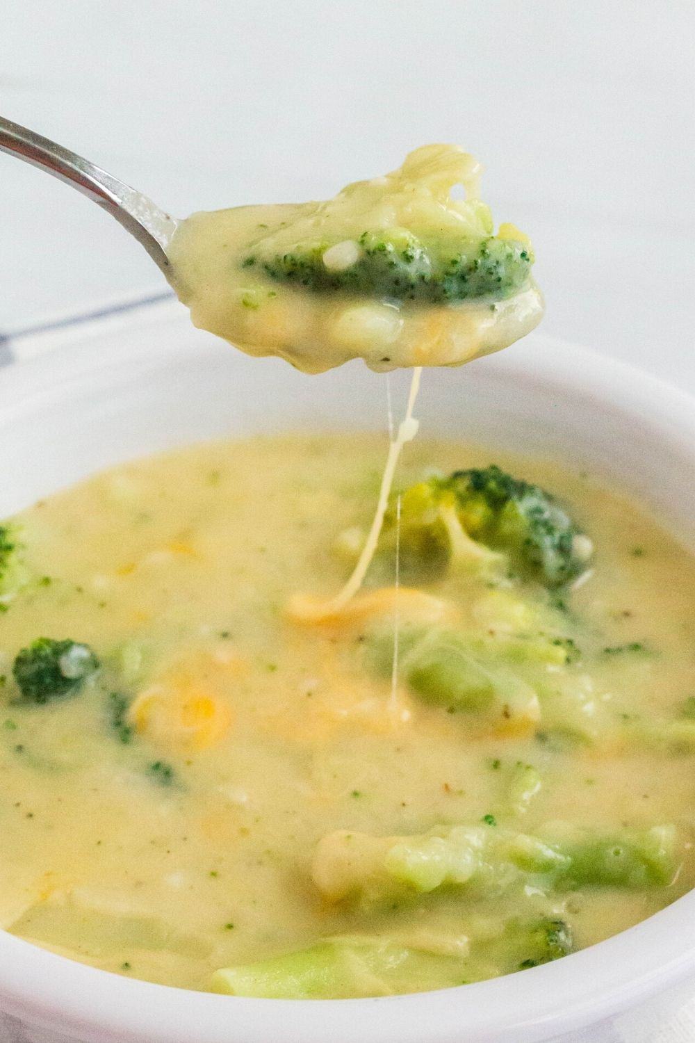 a spoon lifts a bite of Instant Pot potato and broccoli soup out of a white bowl, while cheese pulls stretch from the bowl to the spoon