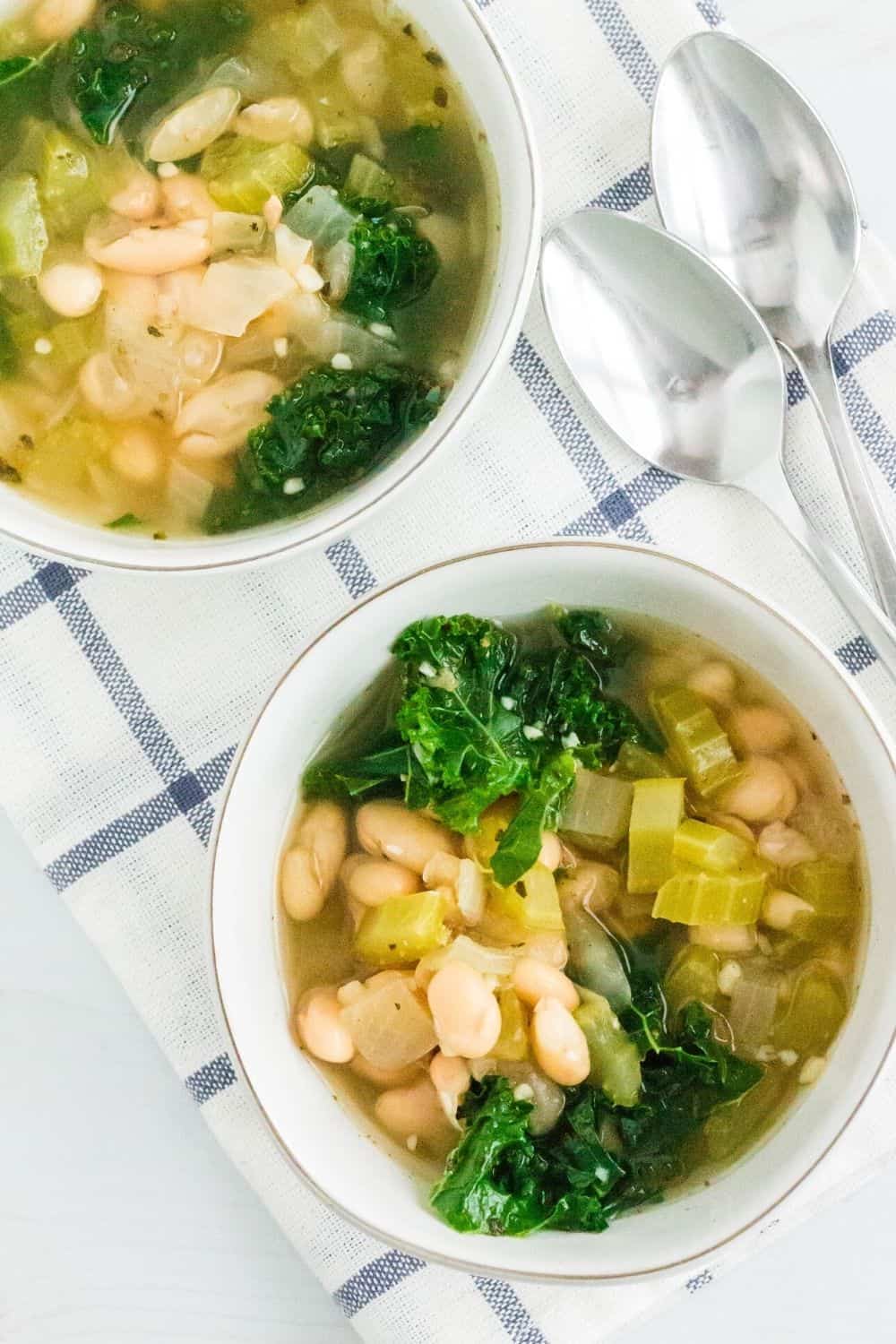 Instant Pot white bean and kale soup served in two white bowls, resting on a blue and white napkin with two spoons nearby.