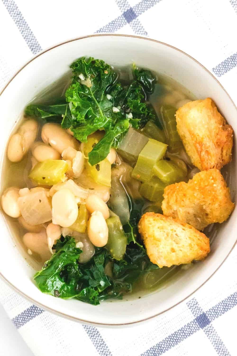 overhead view of a white bowl filled with a serving of white bean and kale soup, topped with three croutons.