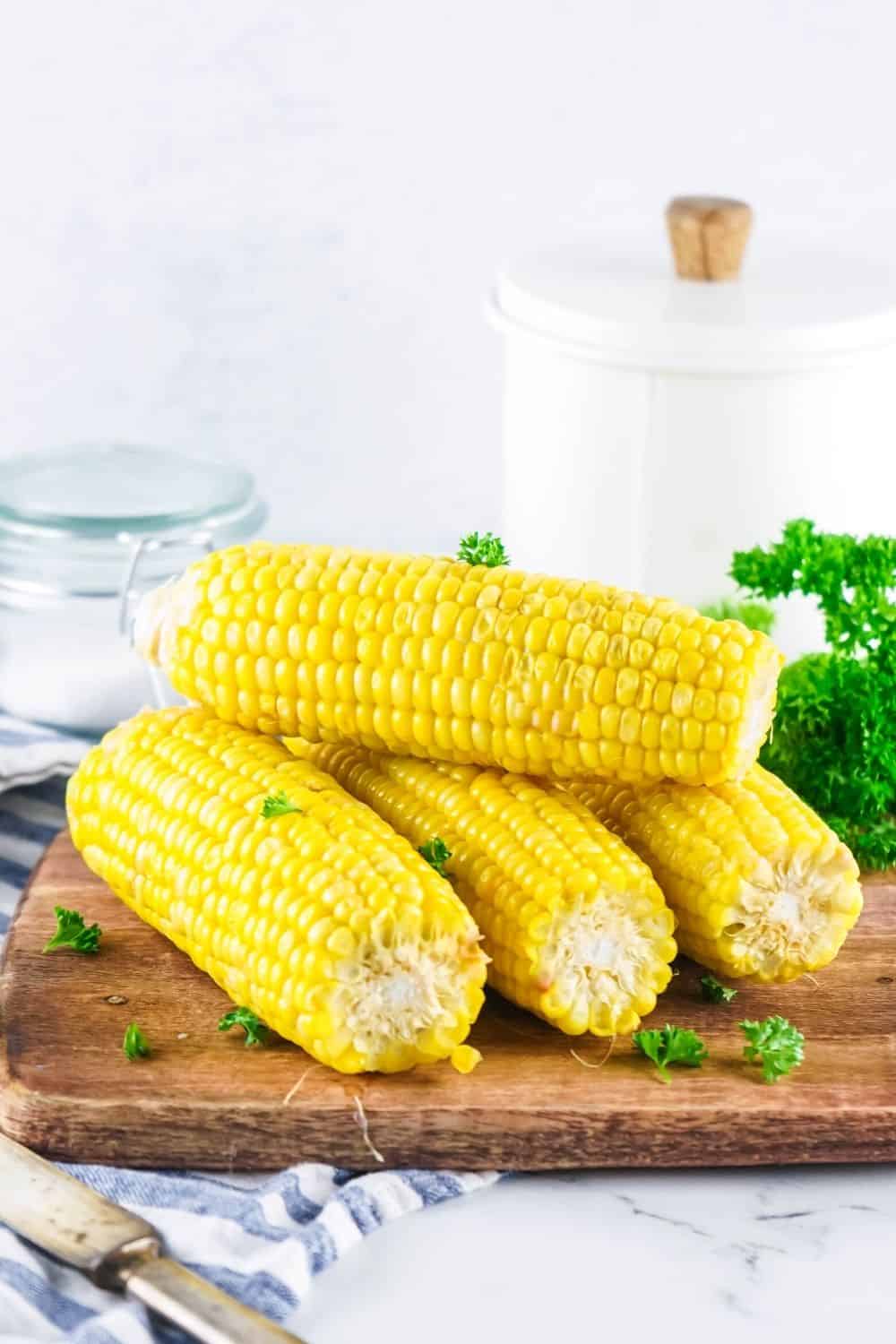 four ears of corn on the cob on a cutting board with parsley and a white canister in the background