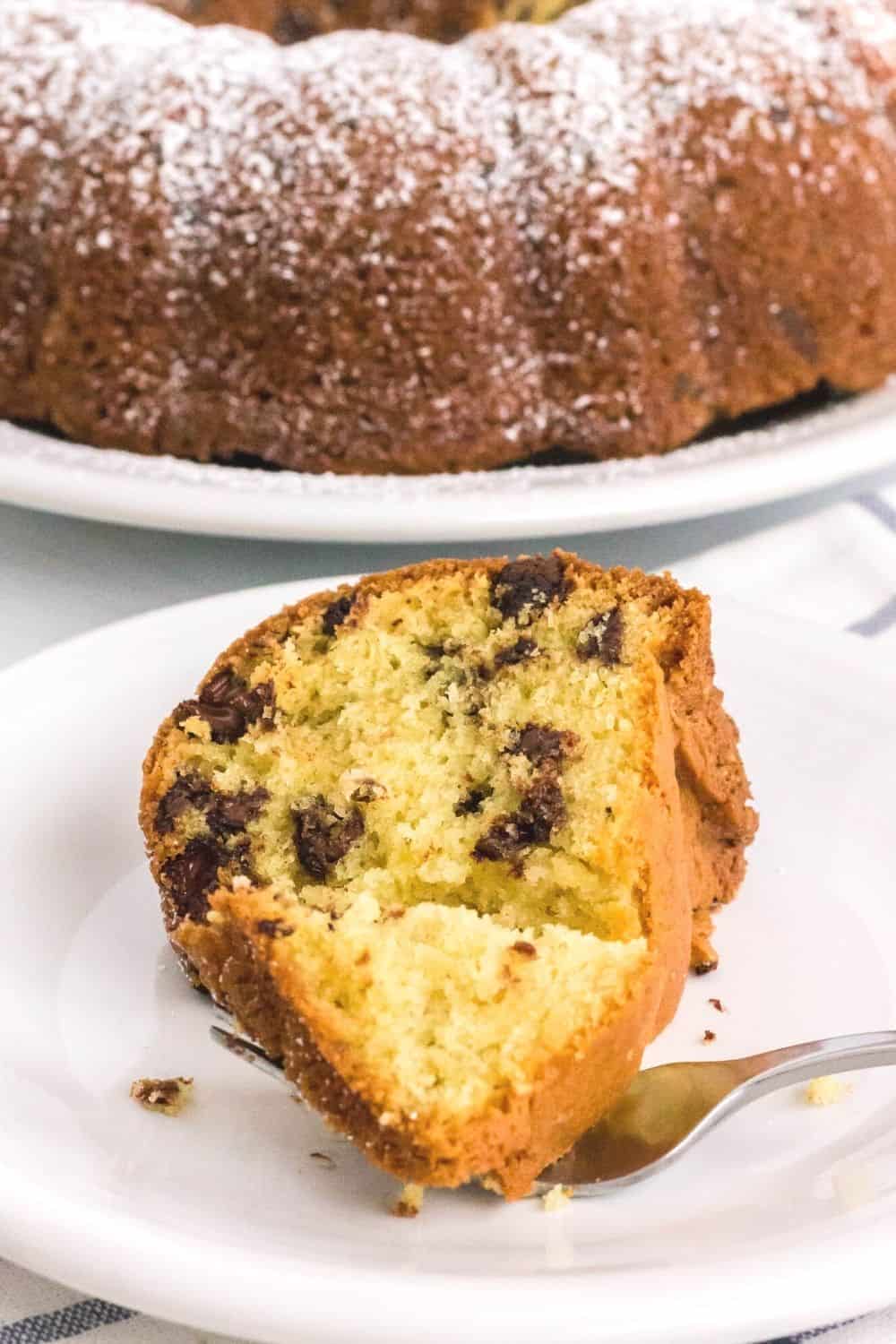 slice of chocolate chip cake on a white plate, with remainder of the bundt cake in the background. A fork has cut a bite out of the slice.