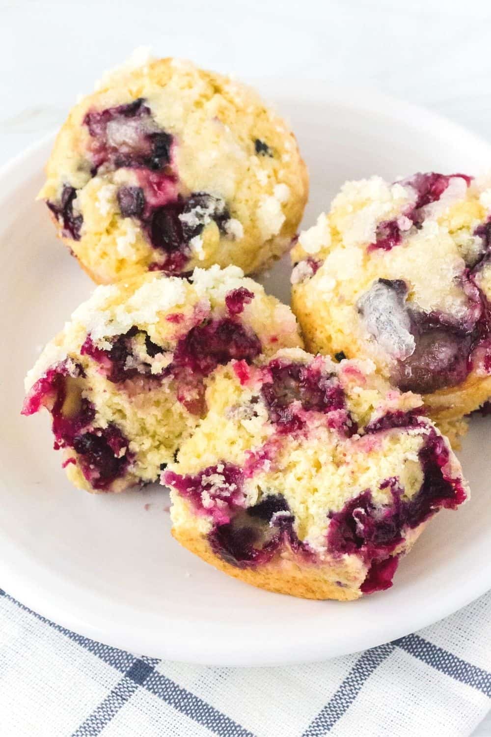 two blueberry streusel muffins on a white plate, along with a third muffin that has been cut in half, showing the burst blueberries inside the tender muffin