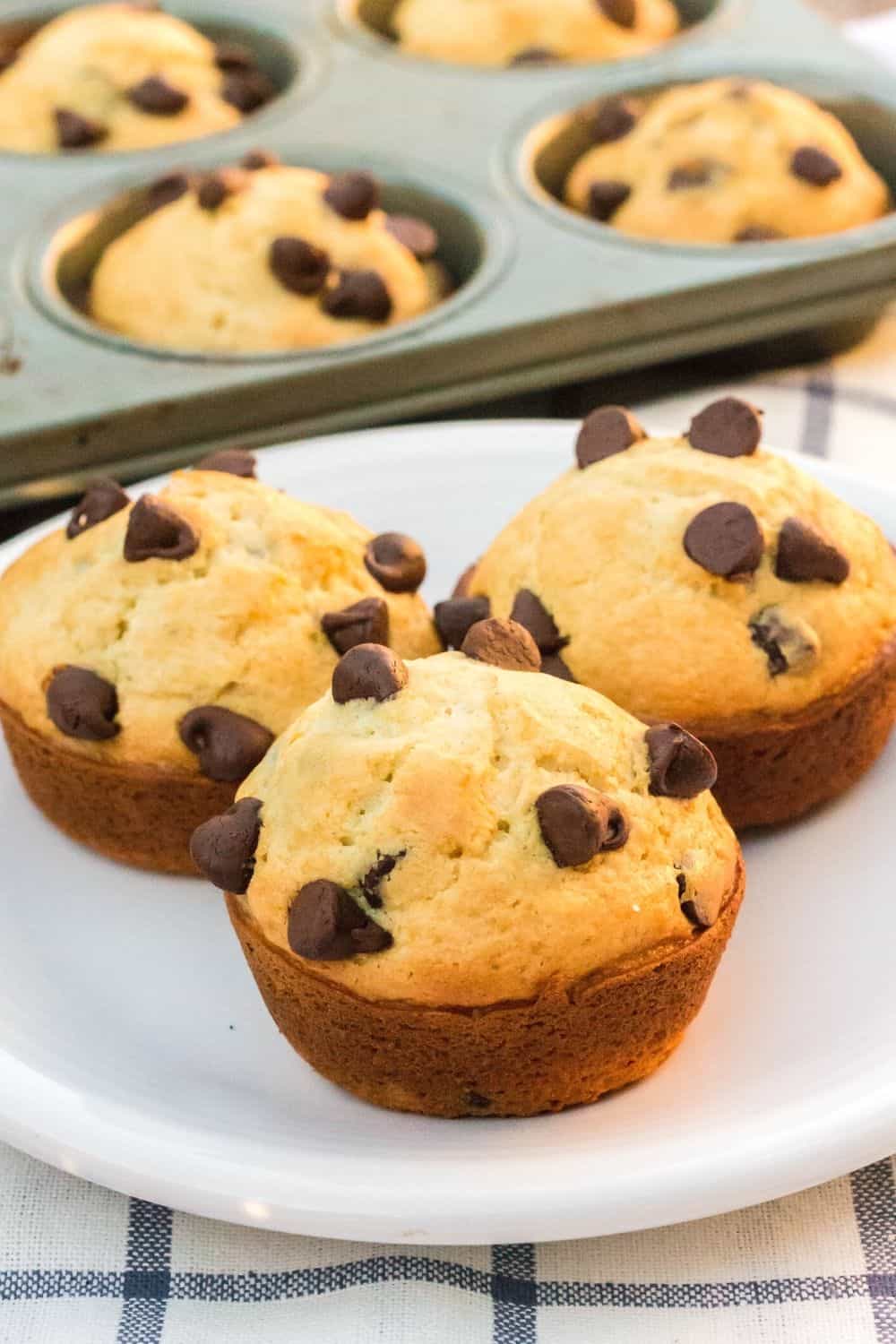 three bisquick chocolate chip muffins on a white plate, with the remainder of the muffins in the background