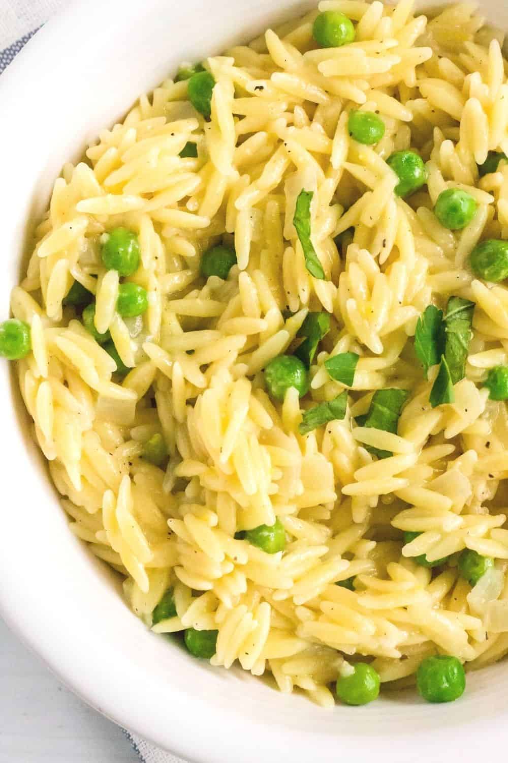 close-up overhead view of a white bowl filled with pressure cooker orzo made with lemon, parmesan, frozen peas, and fresh basil and parsley.