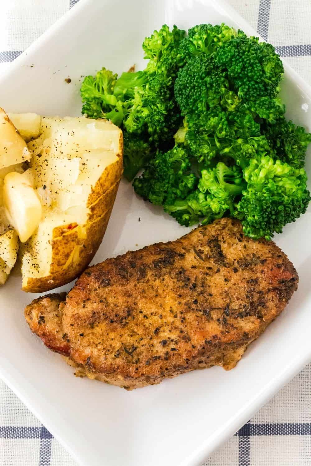 overhead view of a perfectly-seasoned pressure cooker boneless pork chop served on a square white plate with a buttered baked potato and steamed broccoli.