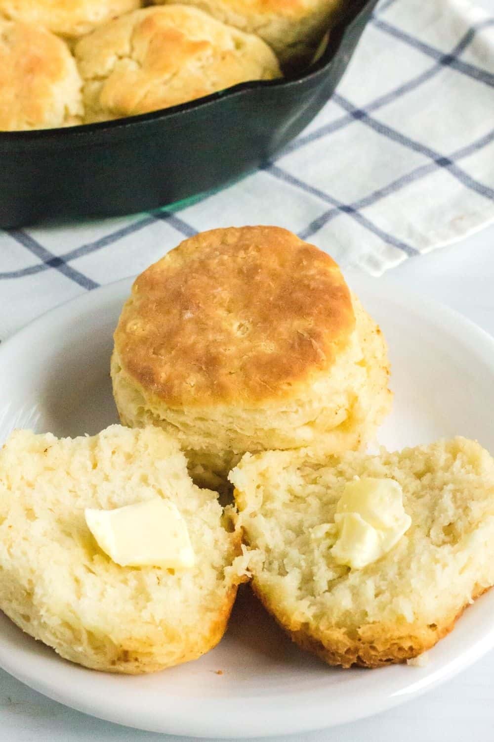 a split biscuit, topped with butter, along with a whole biscuit on a white plate. A cast iron pan of homemade biscuits is in the background.