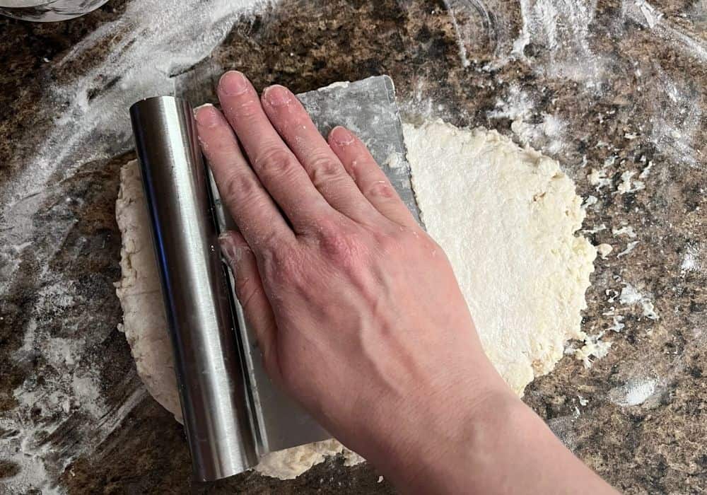 A woman's hand presses on a bench scraper to flatten biscuit dough into a 1-inch disk.