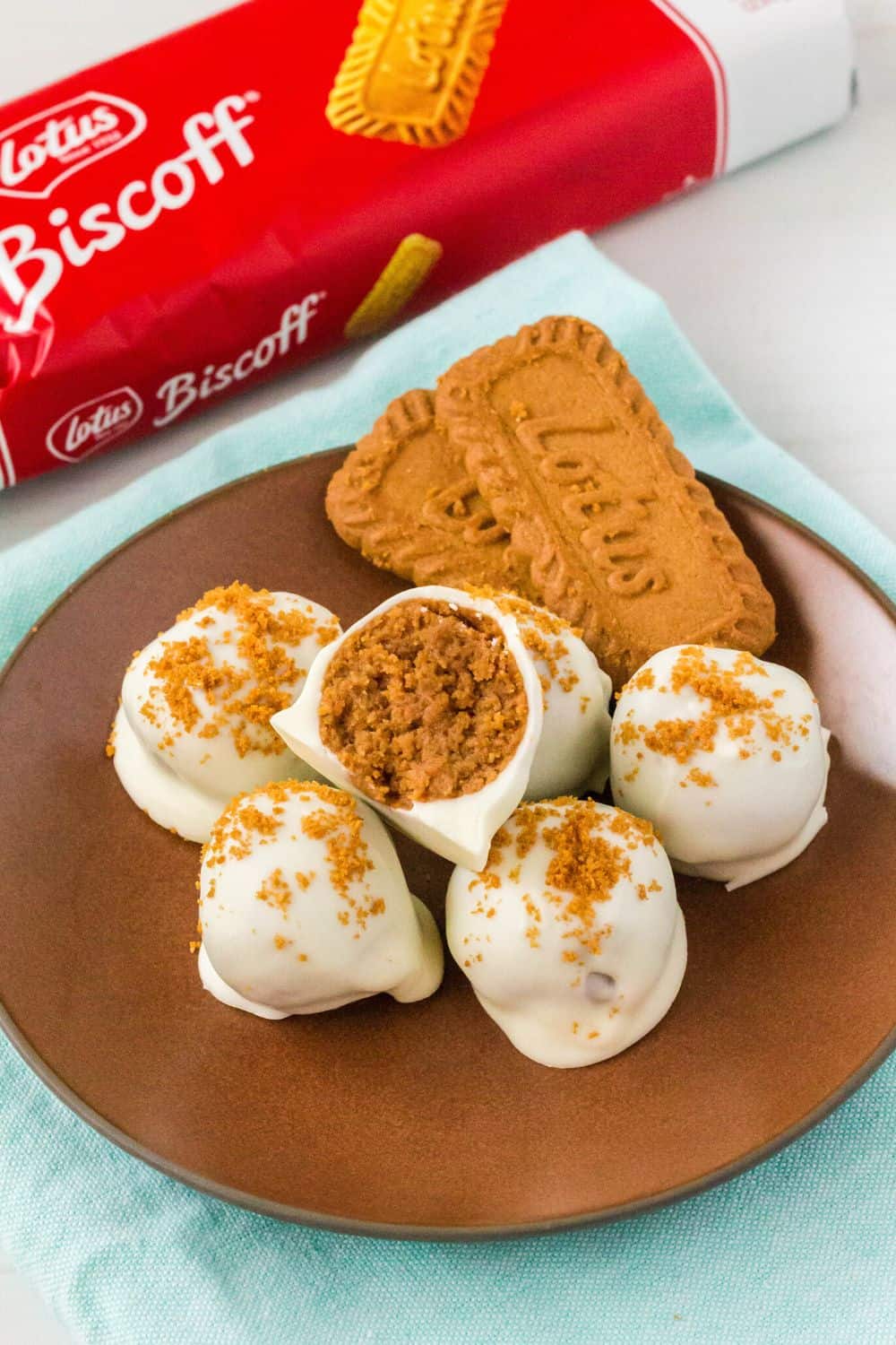 no-bake Biscoff truffles on a brown plate, with one truffle cut in half to show the interior. Two lotus cookies are on the plate, with the package of Biscoff cookies in the background.