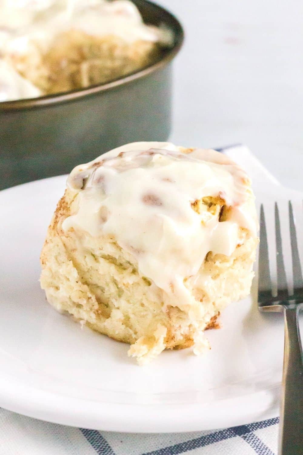 fluffy homemade cinnamon roll on a white plate with a fork next to it. The pan of cinnamon rolls is in the background.