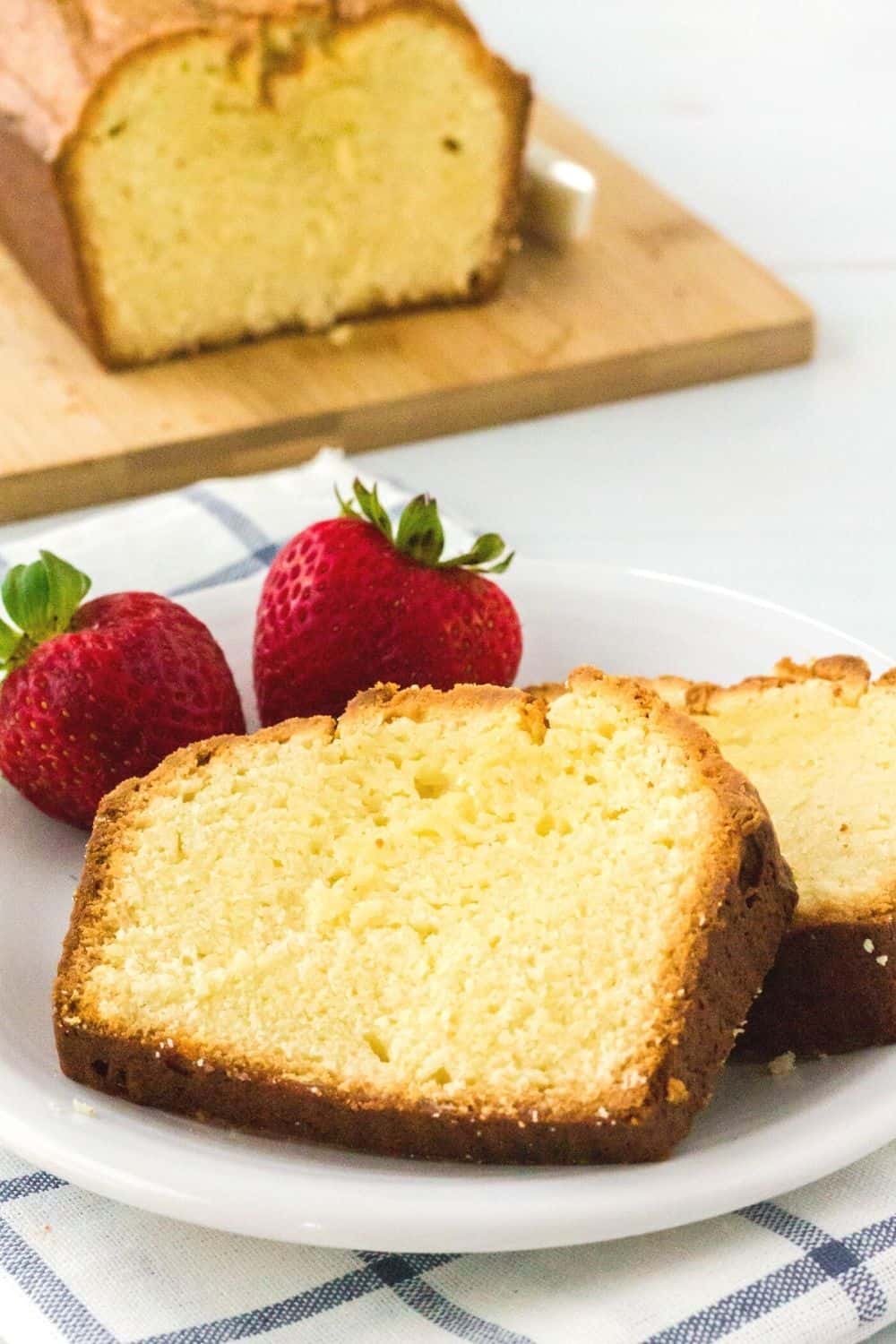 two slices of cream cheese pound cake on a white plate with two strawberries, and the remaining loaf of pound cake on a cutting board in the background