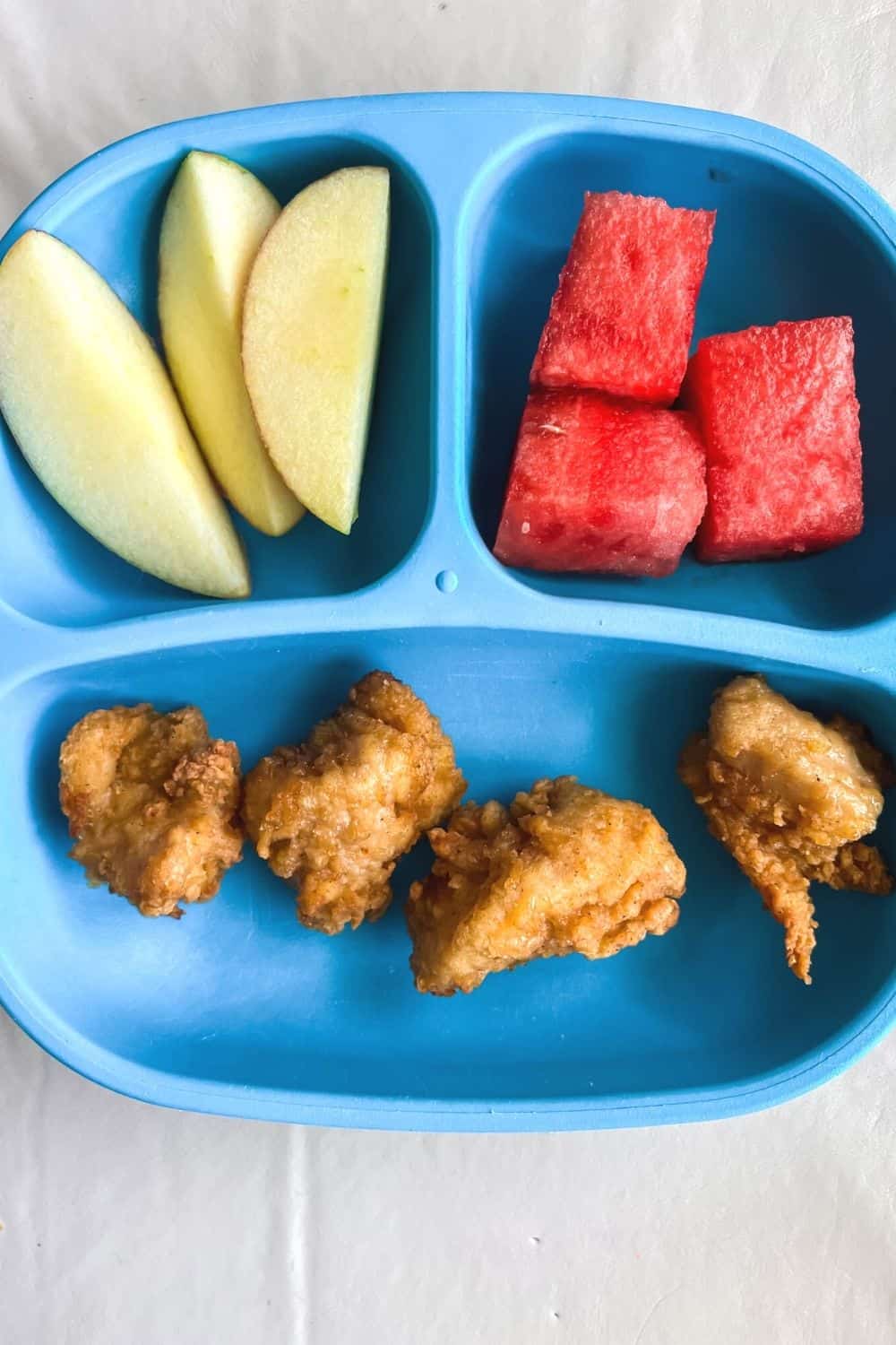 blue divided plastic plate with air fryer chicken nuggets, apple slices, and watermelon cubes on it.
