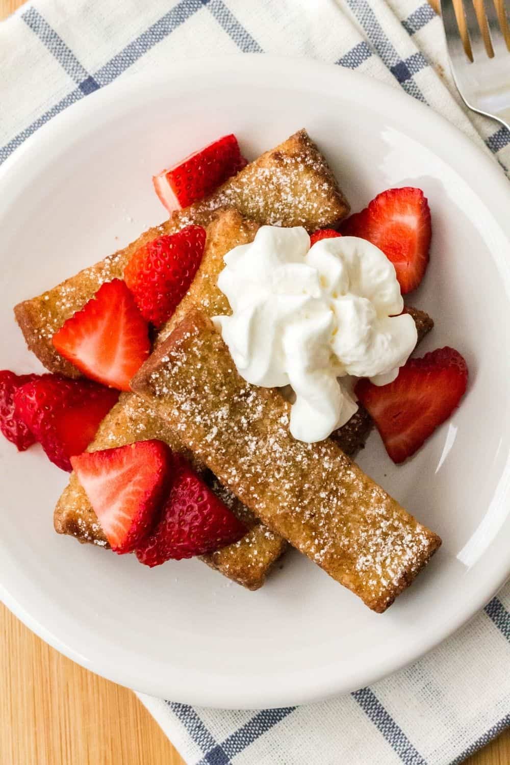white plate serving air fryer frozen french toast sticks with strawberries and whipped cream, served atop a blue and white napkin