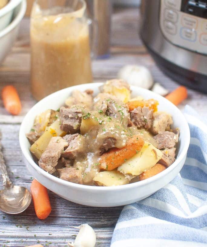 white bowl of Instant Pot pork roast with potatoes and carrots, with gravy in the background