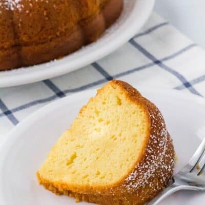 slice of vanilla pudding bundt cake on a white plate, with the remainder of the bundt cake in the background