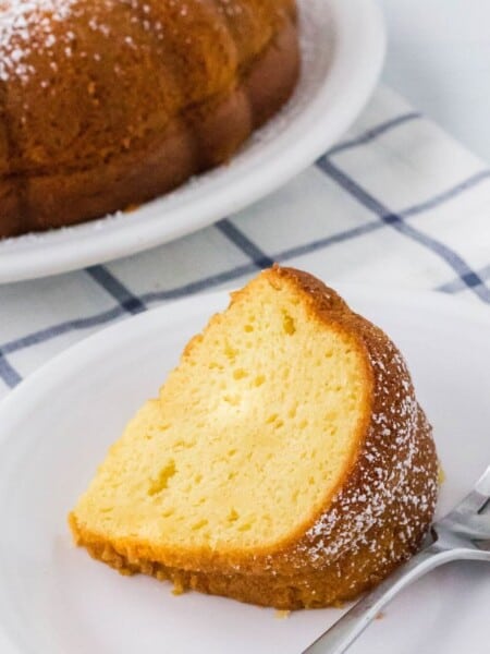 slice of vanilla pudding bundt cake on a white plate, with the remainder of the bundt cake in the background